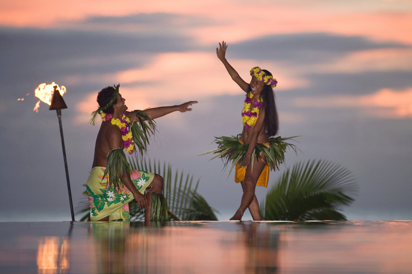 Two performers in traditional Polynesian attire, dancing at sunset with tiki torches in the background.