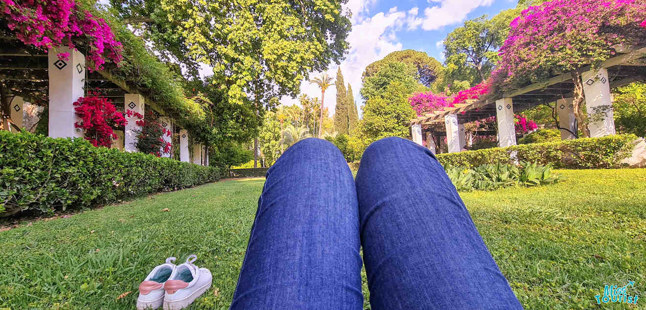 Relaxing perspective of a person sitting on the lush green grass in Maria Luisa Park, Seville, with blooming pink bougainvillea and tall palm trees in the background, and a pair of casual shoes placed on the grass foreground