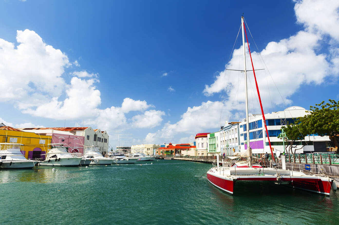 A harbor with sailboats and yachts docked along the waterfront, colorful buildings in the background, under a clear blue sky with scattered clouds.