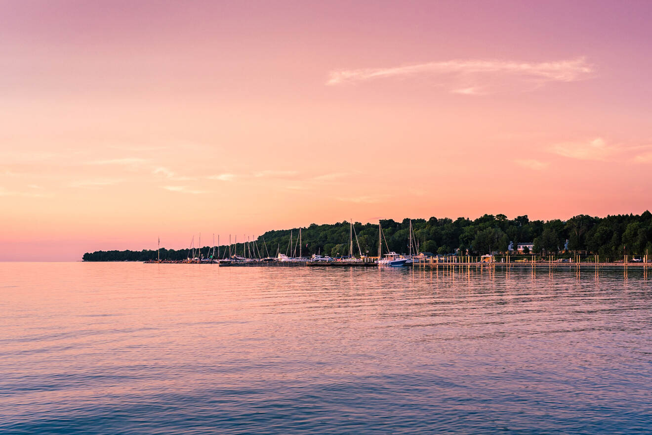 A serene lakeside scene at sunset, with various sailboats docked along the shoreline and a forested area in the background under a pink sky.