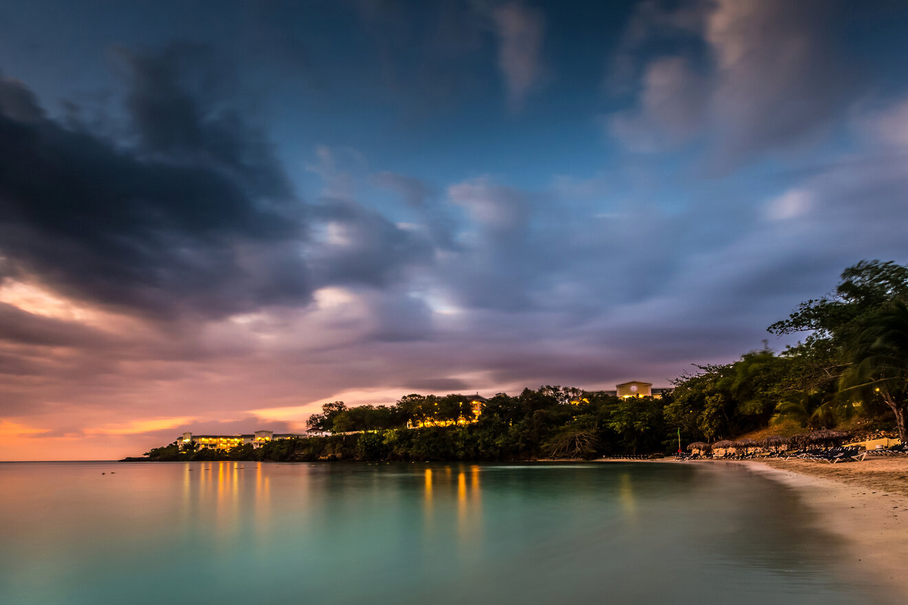 A serene beach scene at sunset with calm waters, a distant shoreline dotted with trees and buildings, and a colorful sky filled with clouds.