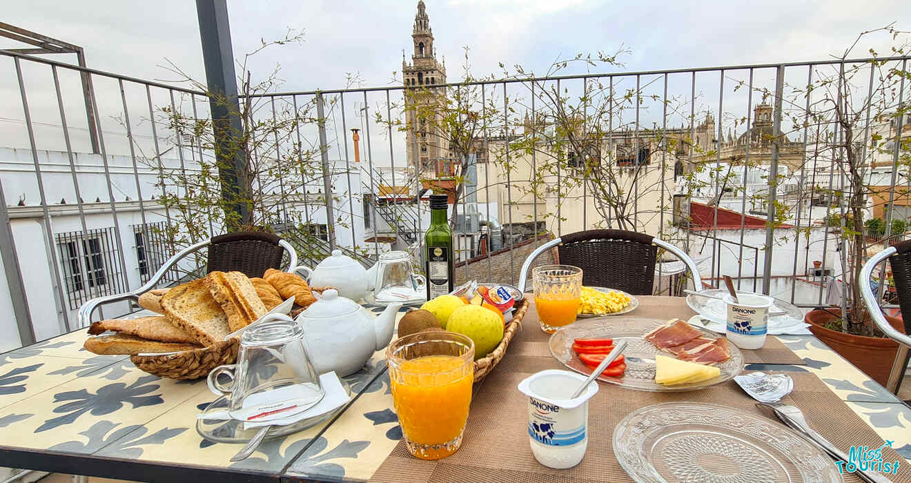 An inviting breakfast spread on a terrace table, complete with fresh fruits, assorted breads, orange juice, and a view of the Giralda Tower in Seville, invites a delightful start to the day