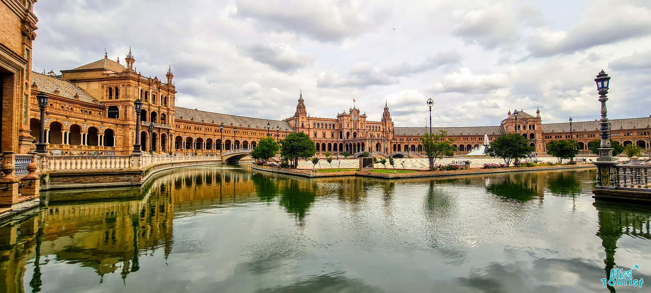 Panoramic view of Plaza de España in Seville with its semi-circular brick building, renaissance towers, and bridges reflecting over the calm canal, under a cloudy sky.