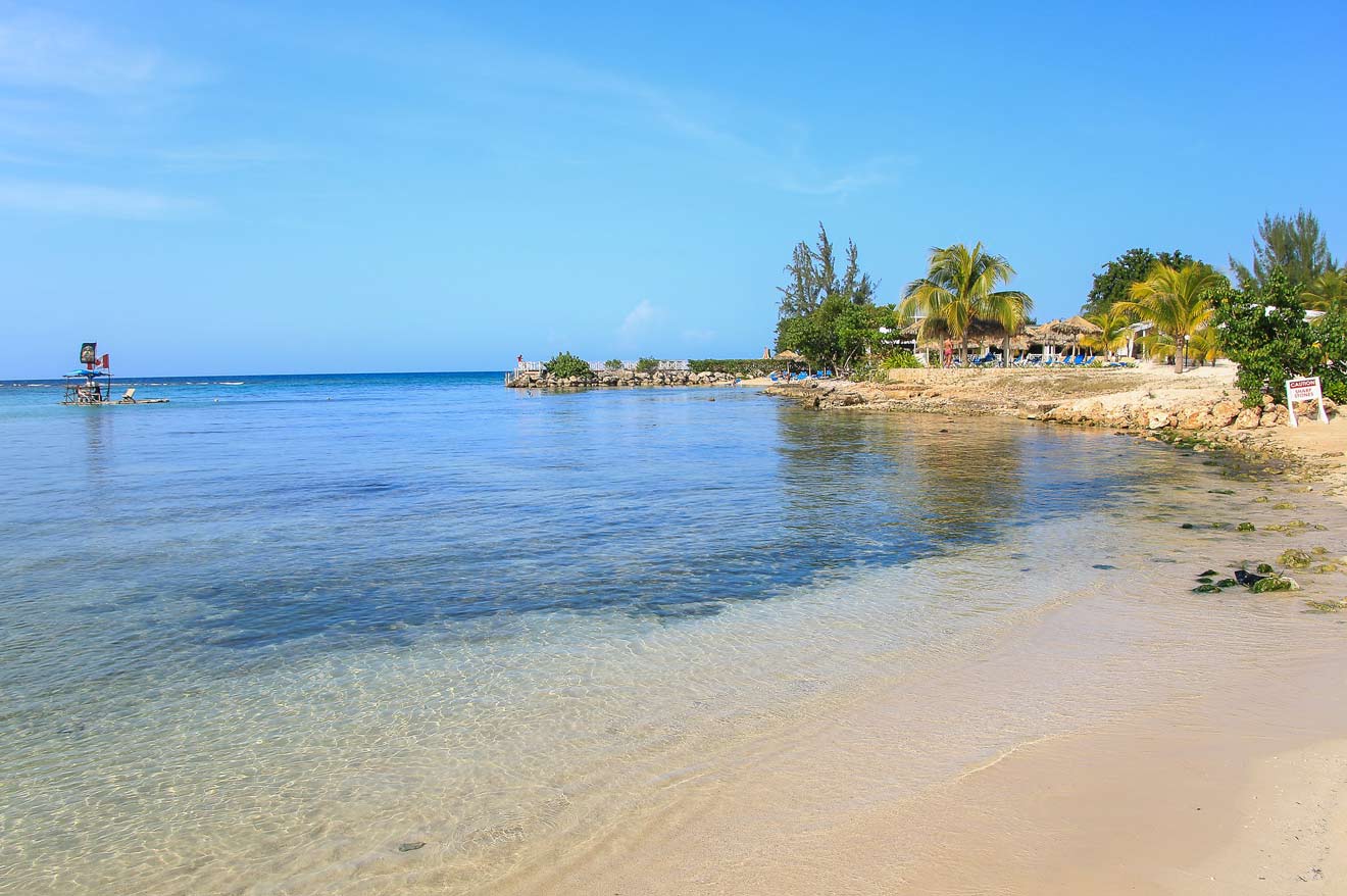 A tranquil beach scene with clear blue water, sandy shoreline, and palm trees. The sky is clear, and there's a lifeguard tower in the distance.