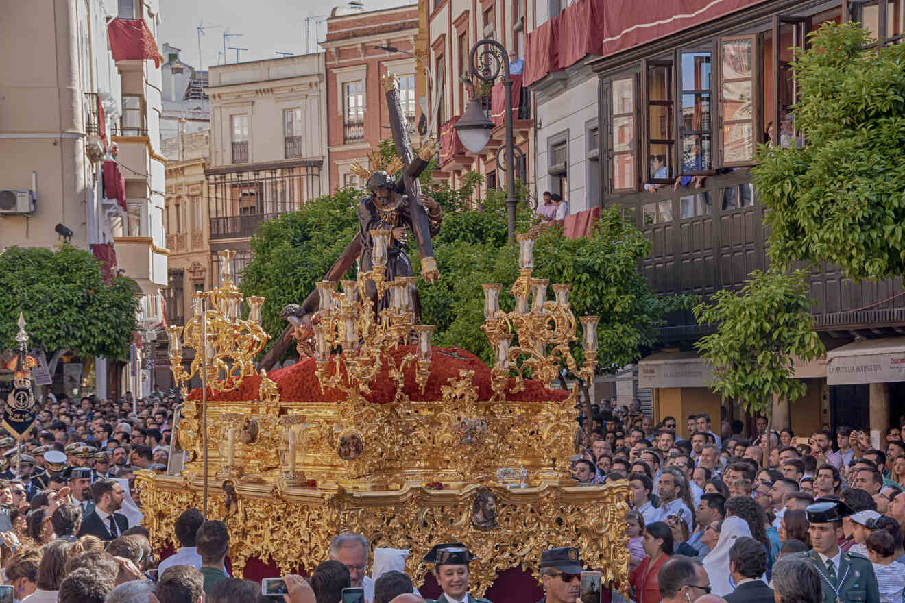 A crowd gathers to witness a traditional Semana Santa procession in Seville, with a highly ornate paso (float) carrying a statue of Jesus amidst gold candlesticks