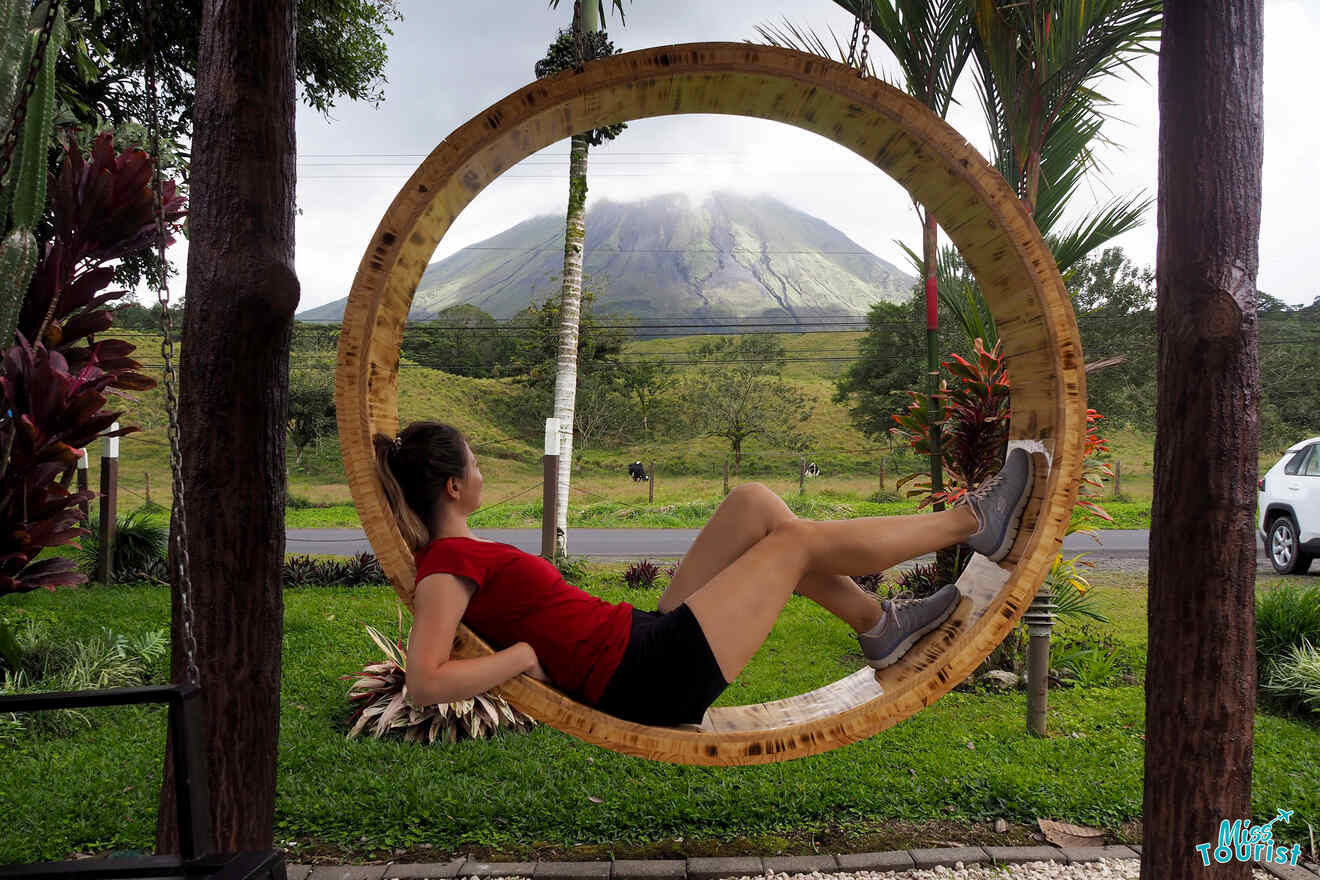 Author of the post in a red shirt and black shorts is sitting in a large wooden ring with a mountain landscape in the background. Trees and plants surround the area.