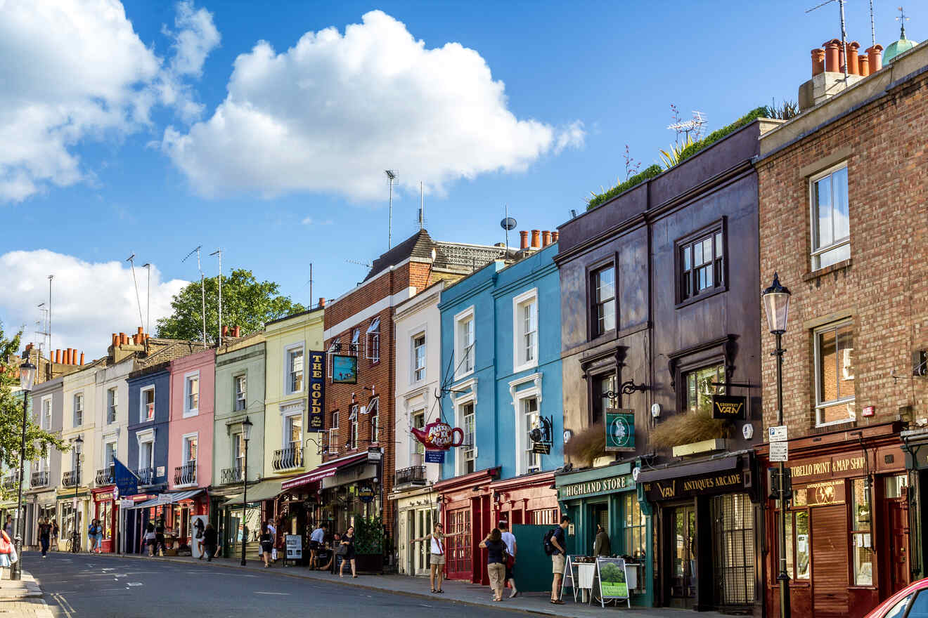 A street with colorful storefronts and buildings in vibrant hues of blue, yellow, red, and brown. People walk along the sidewalk on a bright day with blue skies and scattered white clouds.