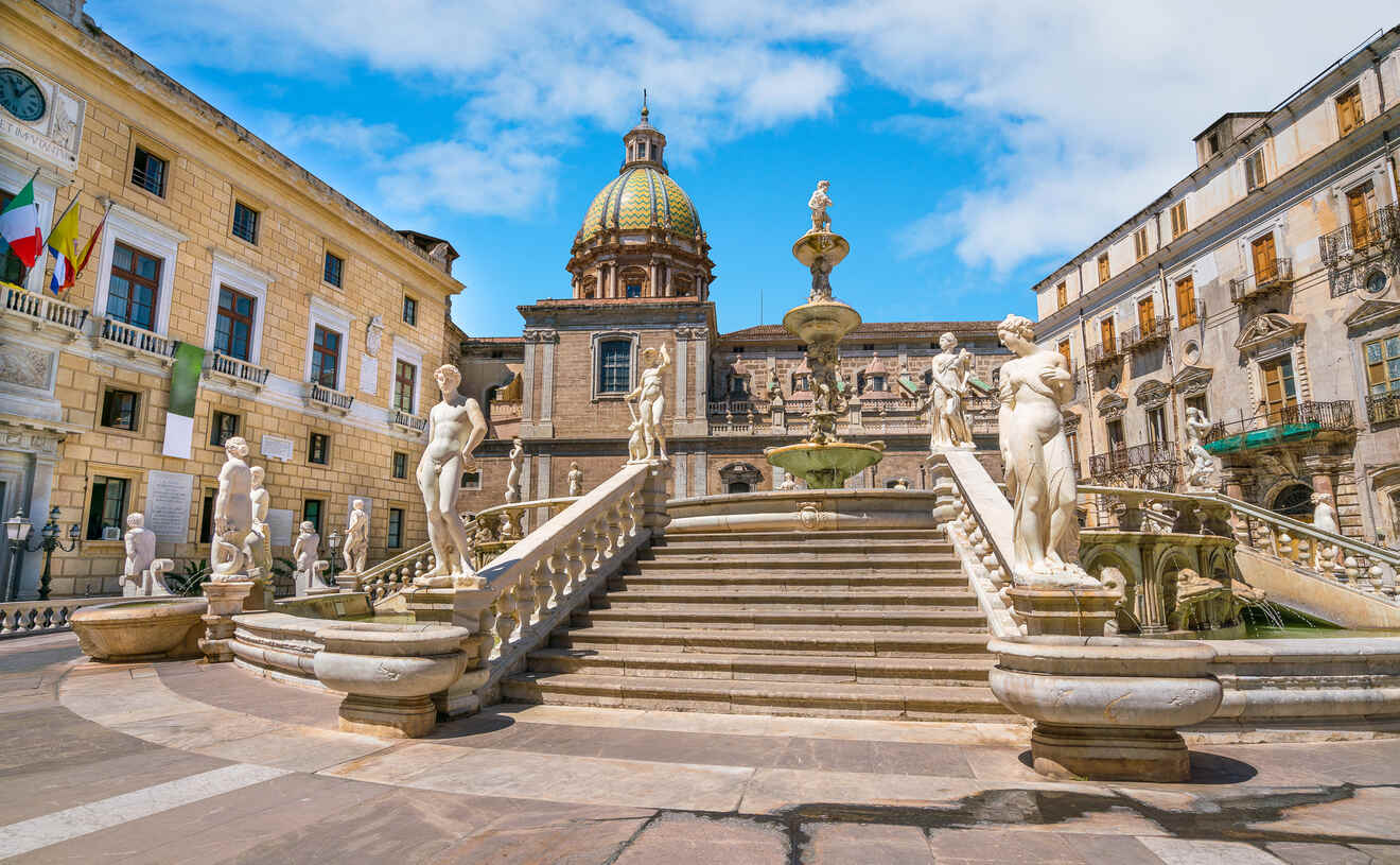 A historical square featuring a baroque fountain, surrounded by ornate statues and steps, with domed buildings and Italian flags in the background under a blue sky.