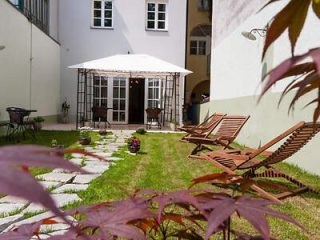 A small, neatly maintained backyard with lounge chairs, potted plants, and a white canopy in front of a building with white walls and multiple windows.