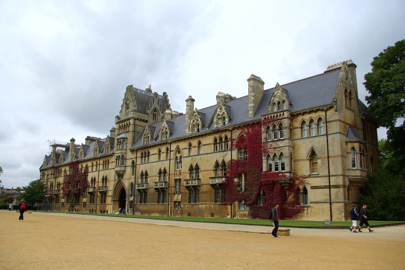 A large, historic building with ivy-covered walls, gothic architecture, and a clock tower. People walk along a gravel path in front of it under a cloudy sky.