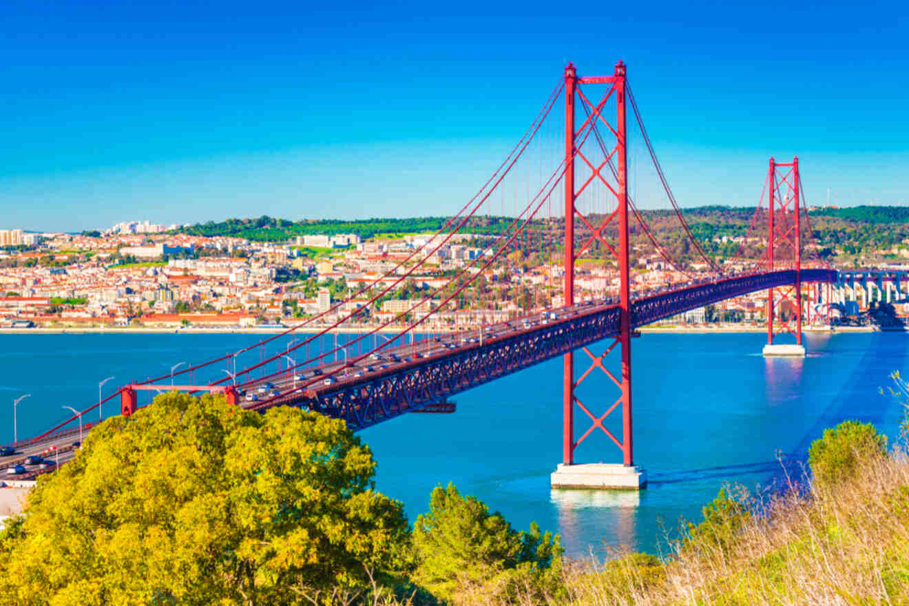 A view of the 25 de Abril Bridge spanning the Tagus River in Lisbon, with the cityscape in the background and lush greenery in the foreground.