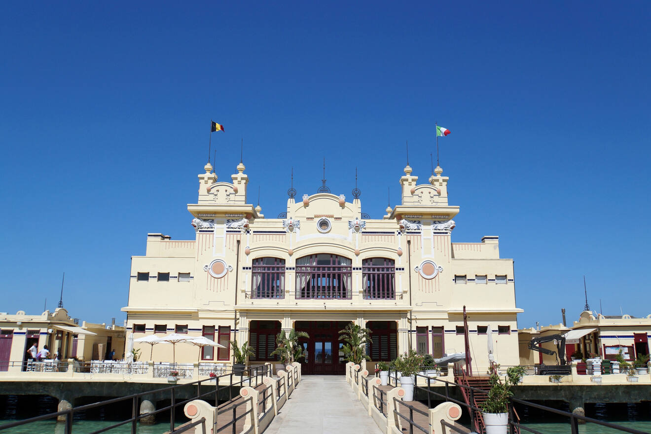 A historic building with ornate architecture under a clear blue sky, featuring a walkway leading up to the entrance. Two flags are flying on top of the structure.
