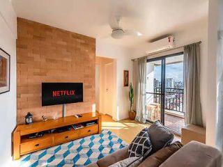 A living room with a mounted TV displaying the Netflix logo, a wooden entertainment unit, a blue and white rug, a sofa, and a sliding glass door leading to a balcony with outdoor seating.