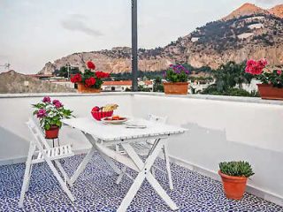 Outdoor patio with a white table and chairs, set with a basket of fruit and plates. Potted flowers decorate the space. Mountains are visible in the background.