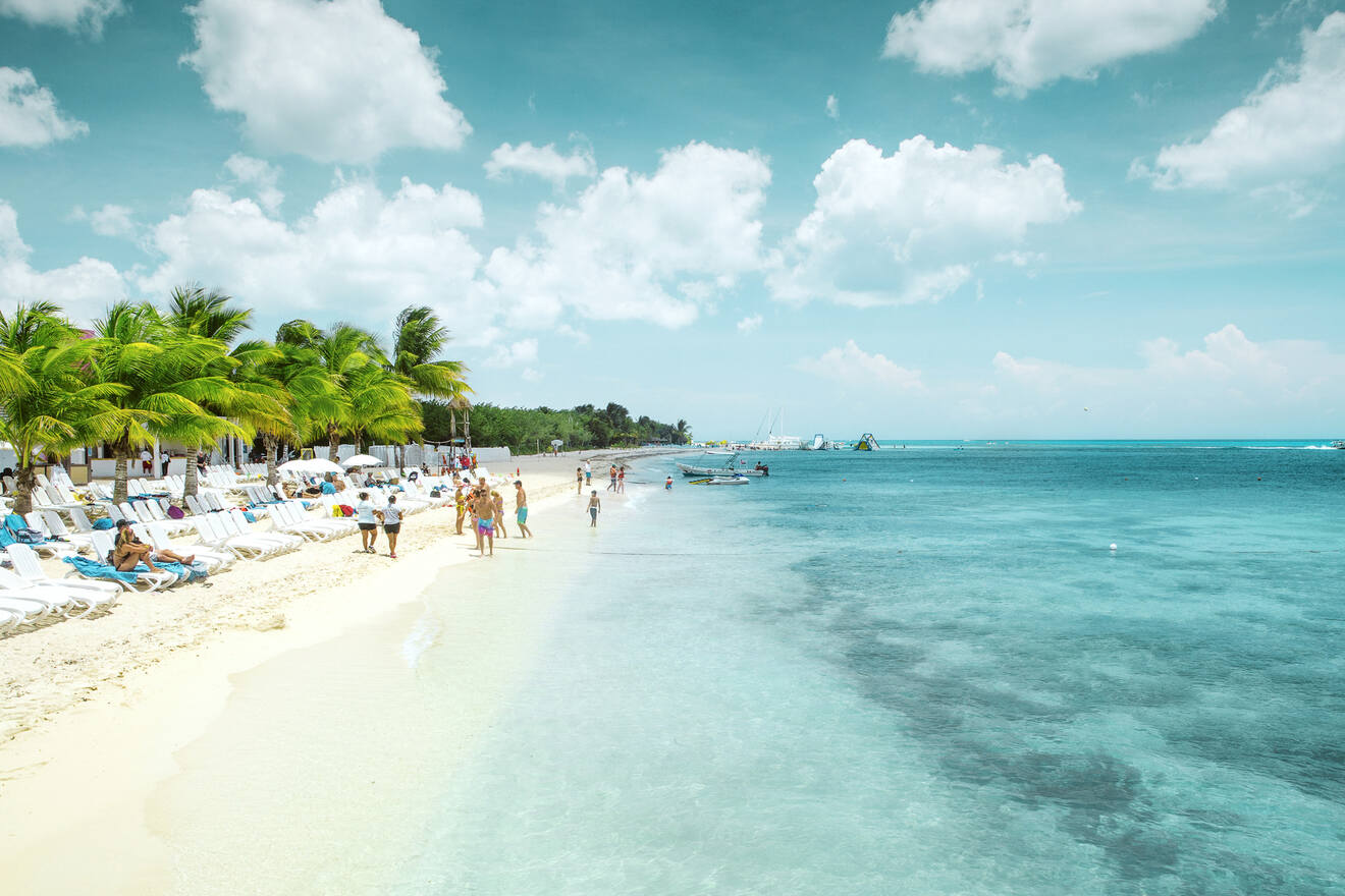 Beach scene with people walking, lounging under palm trees, and boats on clear blue water.