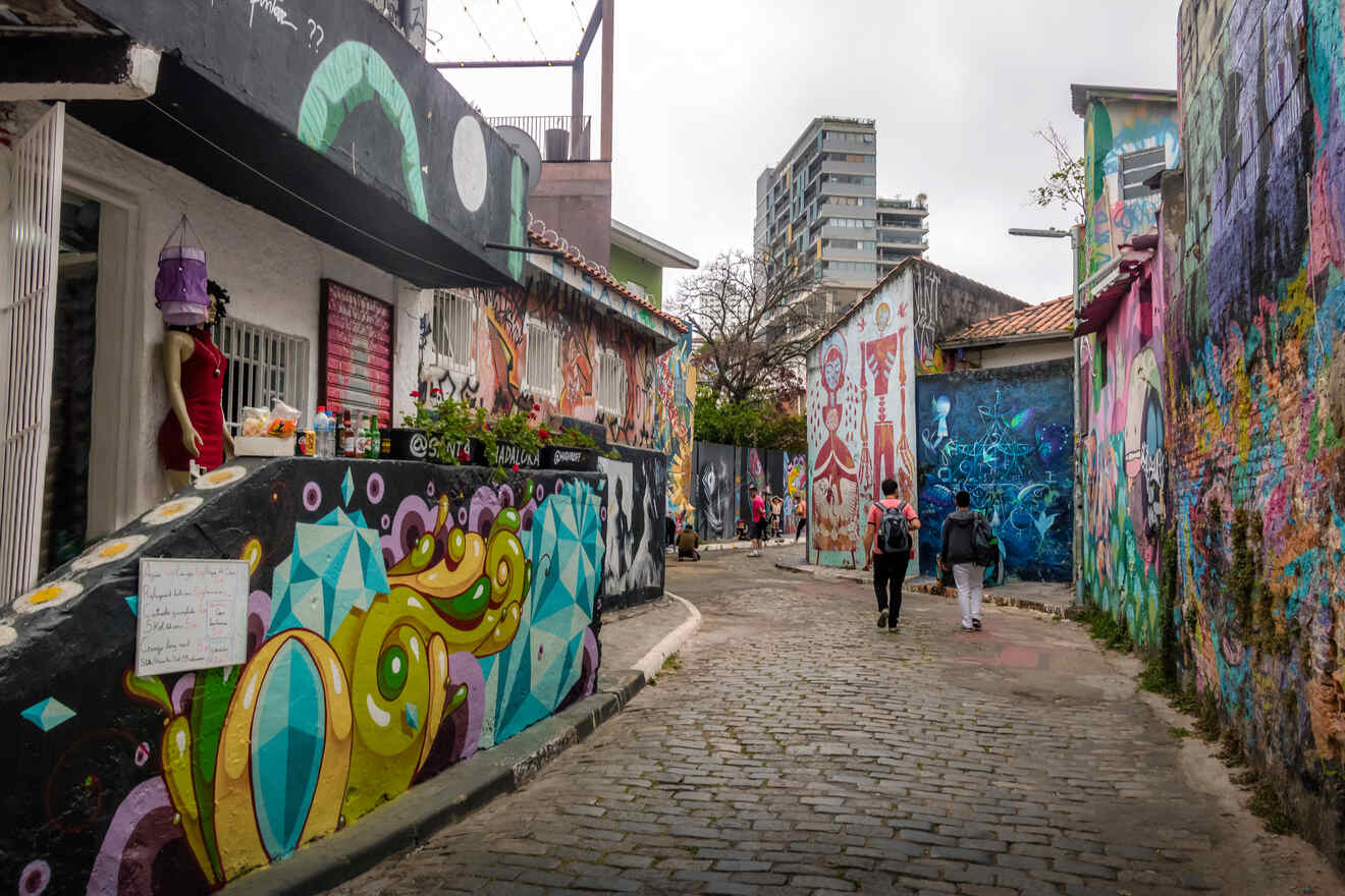 An urban alley features colorful murals and graffiti on building walls. Four people walk along the cobblestone path between the painted walls. City buildings are in the distance.