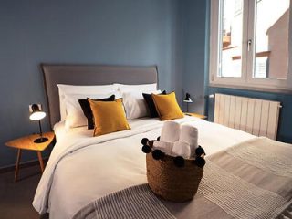 A neatly made double bed with white linens, black and yellow pillows, and a basket of towels on the foot. Nightstands with lamps are on each side, and a window in the background lets in natural light.
