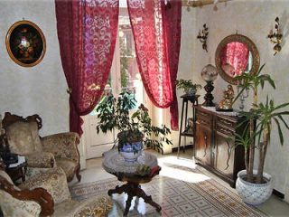 A vintage living room featuring ornate furniture, red lace curtains, potted plants, a wooden cabinet with a circular mirror, and a small table with a decorative plant.