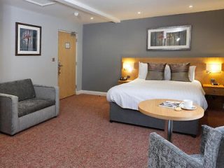 Hotel room with a large bed, two gray armchairs, a round table, a framed picture on the wall, and carpeted floor. A door is in the background.