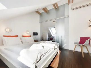 A bright bedroom with a double bed, white linens, and two triangular shaped orange pillows. There is a glass partition on the right leading to another room. A chair with a red blanket is in the corner.