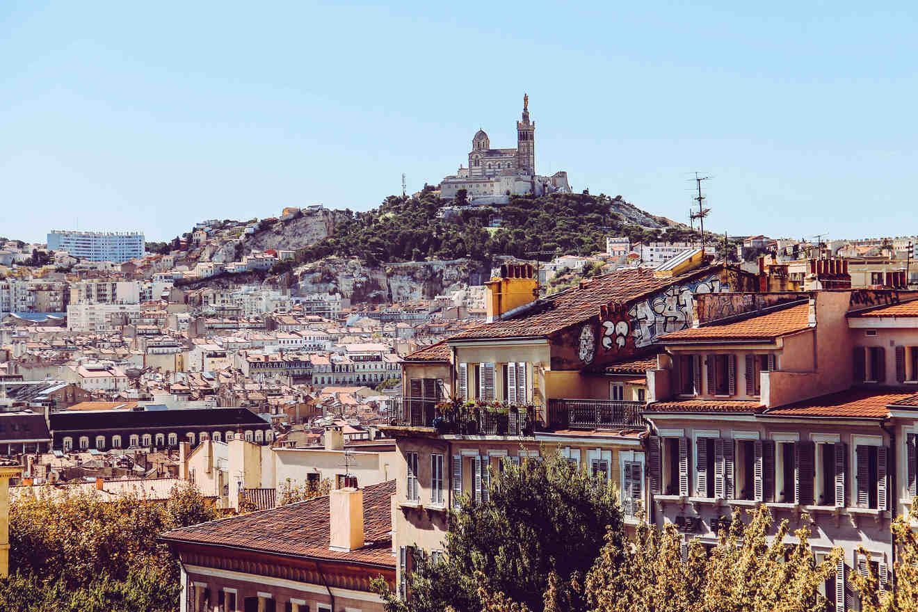 A picturesque view of Marseille cityscape with Notre-Dame de la Garde basilica perched on a hilltop, as seen from a vantage point amidst urban buildings.