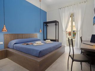 A modern bedroom with a blue accent wall features a bed with blue bedding, two hanging lamps, a small desk and chair, a coat rack, and a window with sheer curtains allowing natural light in.