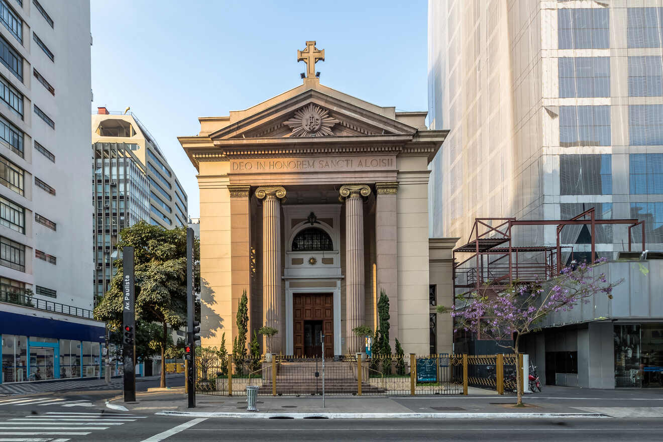 A classical-style church with a prominent entrance and a cross at the top, situated between modern buildings on a city street. The church has a sign that reads "DEO IN HONOREM SANCTI ALOYSII.