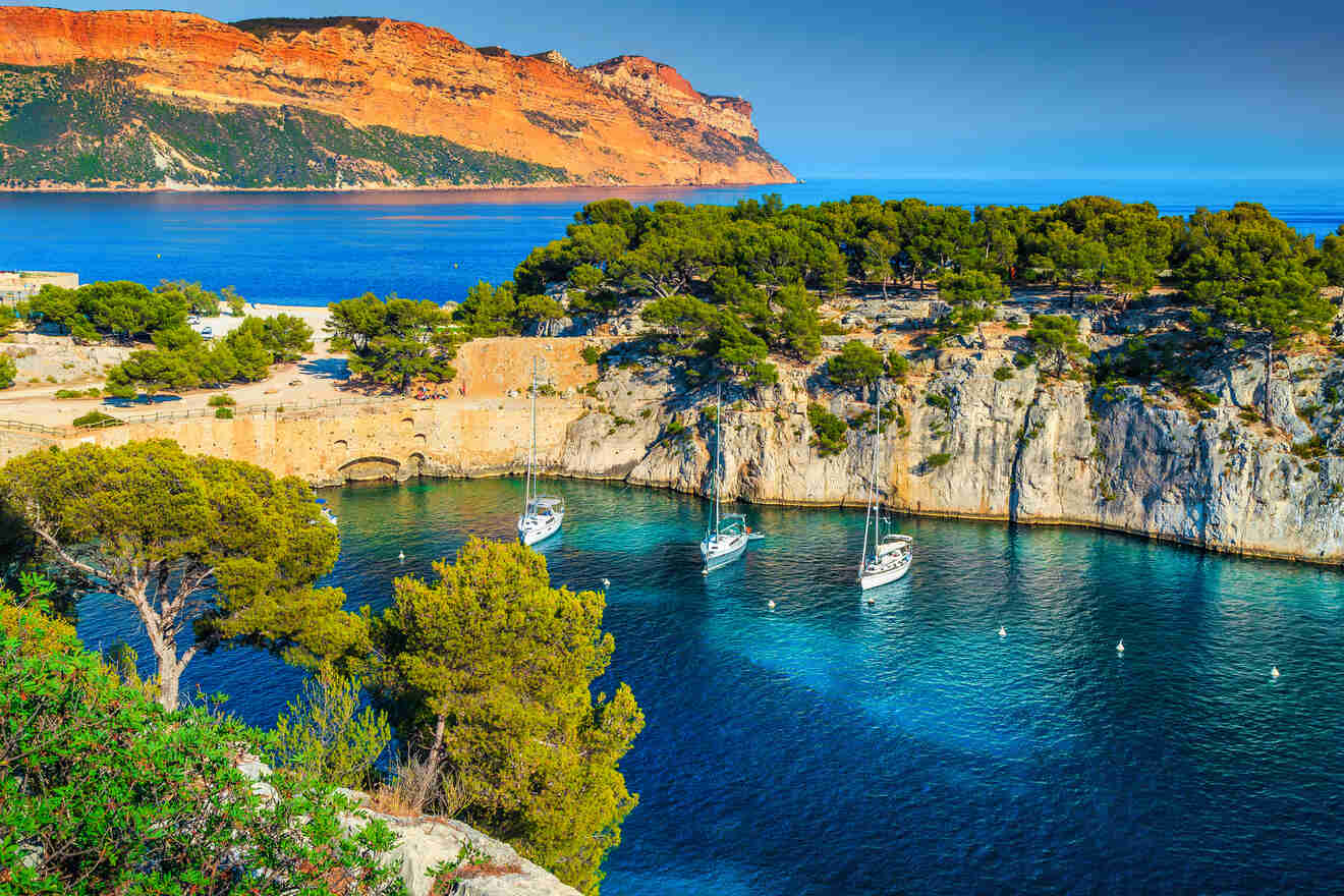 A stunning view of a secluded cove with turquoise waters, anchored sailing boats, surrounded by cliffs and a striking orange mountain backdrop.