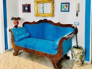 A blue upholstered vintage sofa with wooden carvings sits against a wall with framed artwork, a mirror, a wall-mounted phone, and a decorative vase on the floor beside the sofa.