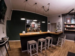 A modern bar setup in a residential space with wooden countertops, four bar stools, pendant lighting, wall art, and shelves with books and decor items.