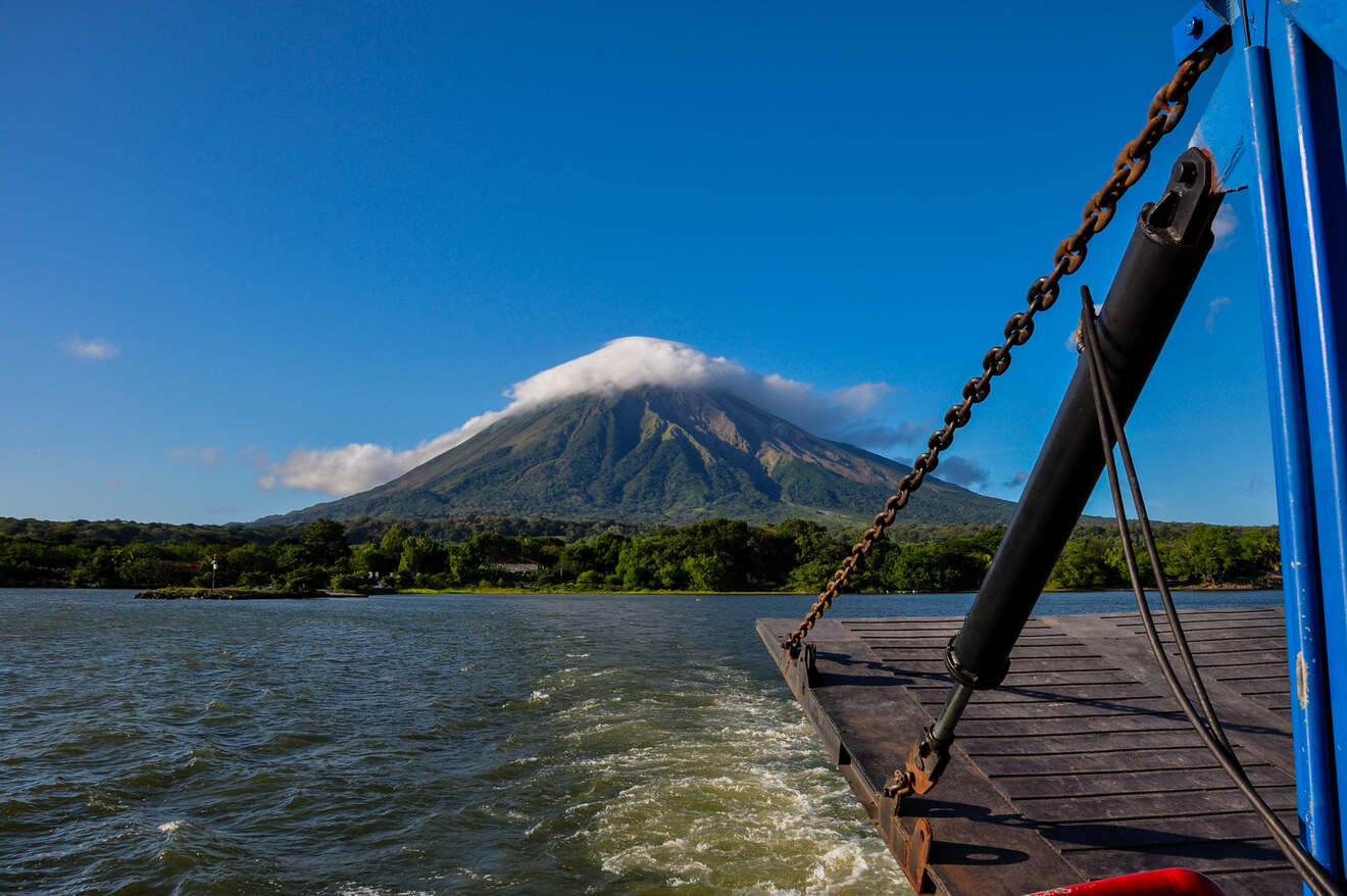 A boat approaches a lush, green volcano with a cloud-covered peak under a clear blue sky.