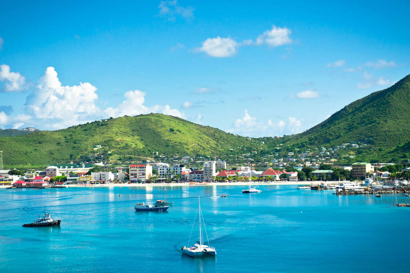 A coastal town with boats moored in bright blue waters, framed by green mountains in the background and a clear blue sky.