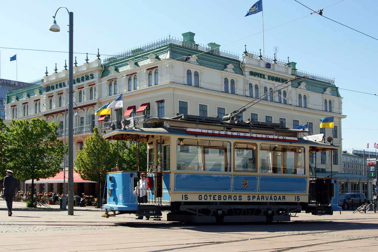 A vintage blue and white tram passes by the Hotel Eggers in Gothenburg, surrounded by trees and flags.