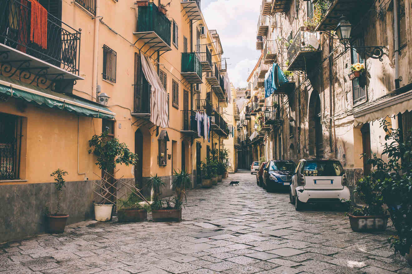 A narrow cobblestone street flanked by yellow buildings with balconies, parked cars, potted plants, and drying laundry under a cloudy sky.