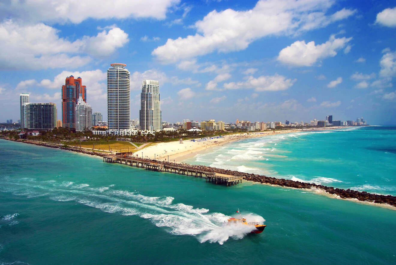 Aerial view of a Miami beach, showing high-rise buildings, a long sandy shoreline, and a speedboat creating waves in the turquoise water.