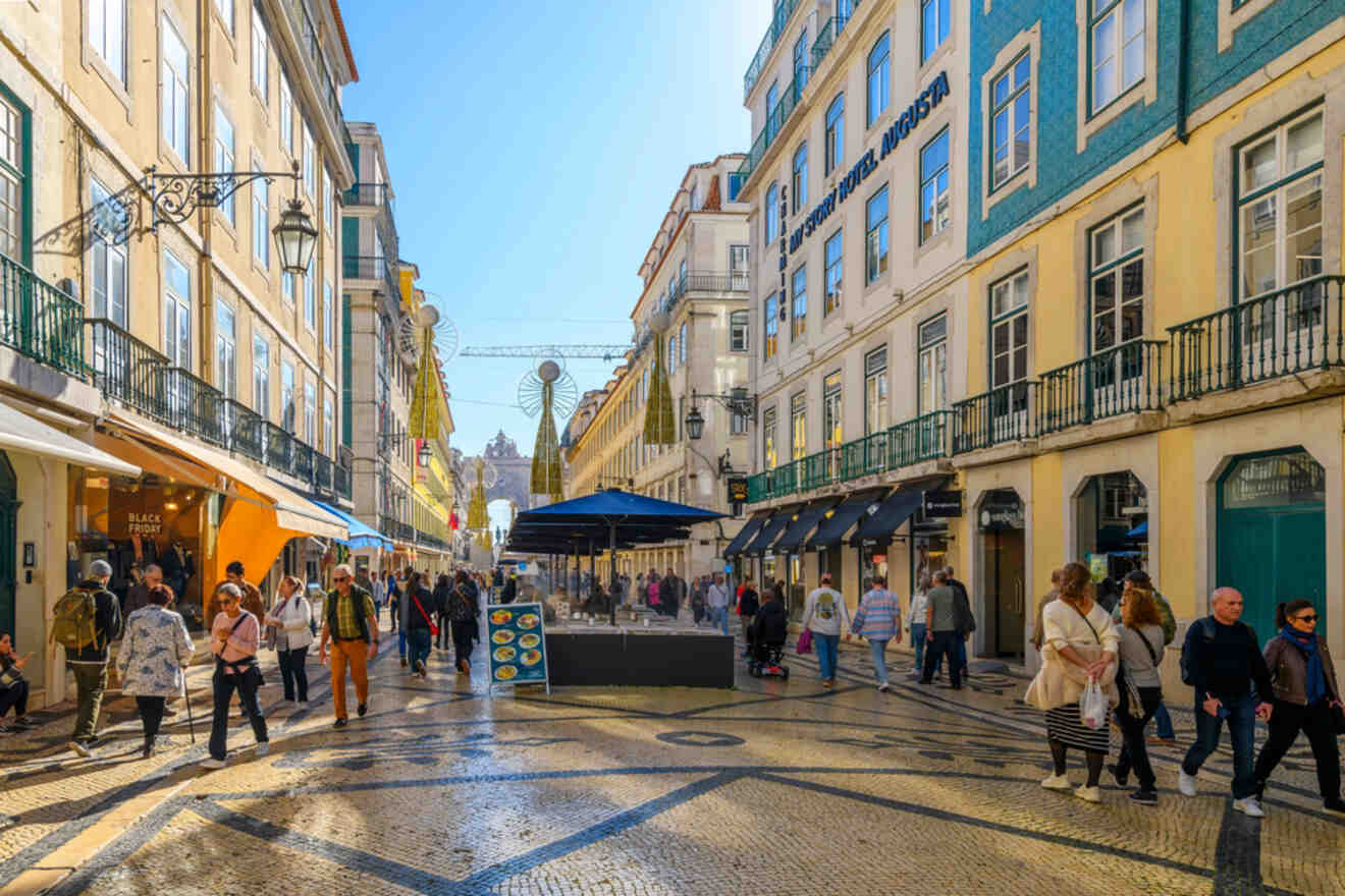 A bustling street in the Bairro Alto district of Lisbon, filled with people walking and shopping, lined with colorful buildings and outdoor cafes under a clear blue sky