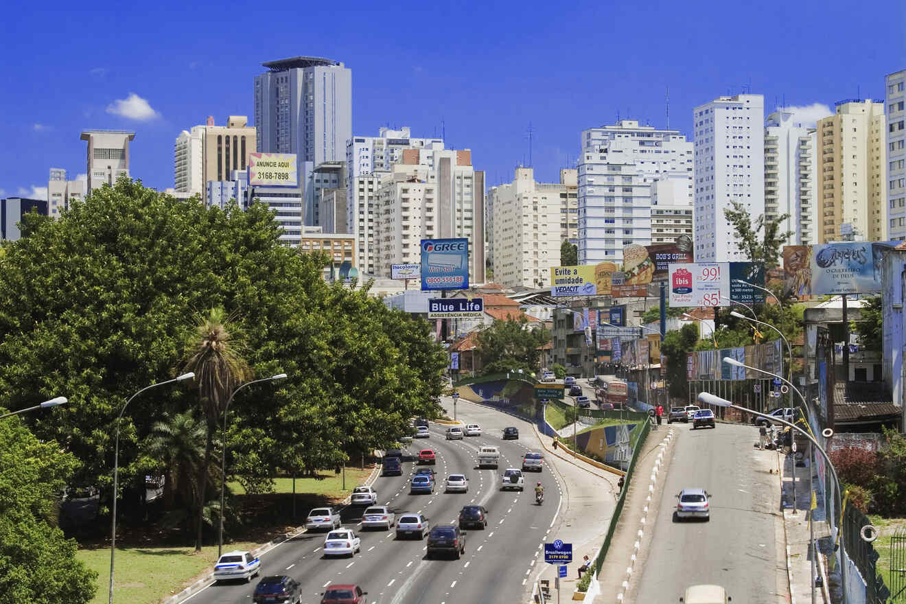 Cityscape featuring a busy road with multiple vehicles, surrounded by high-rise buildings under a clear blue sky. Trees and billboards line the sides of the street.