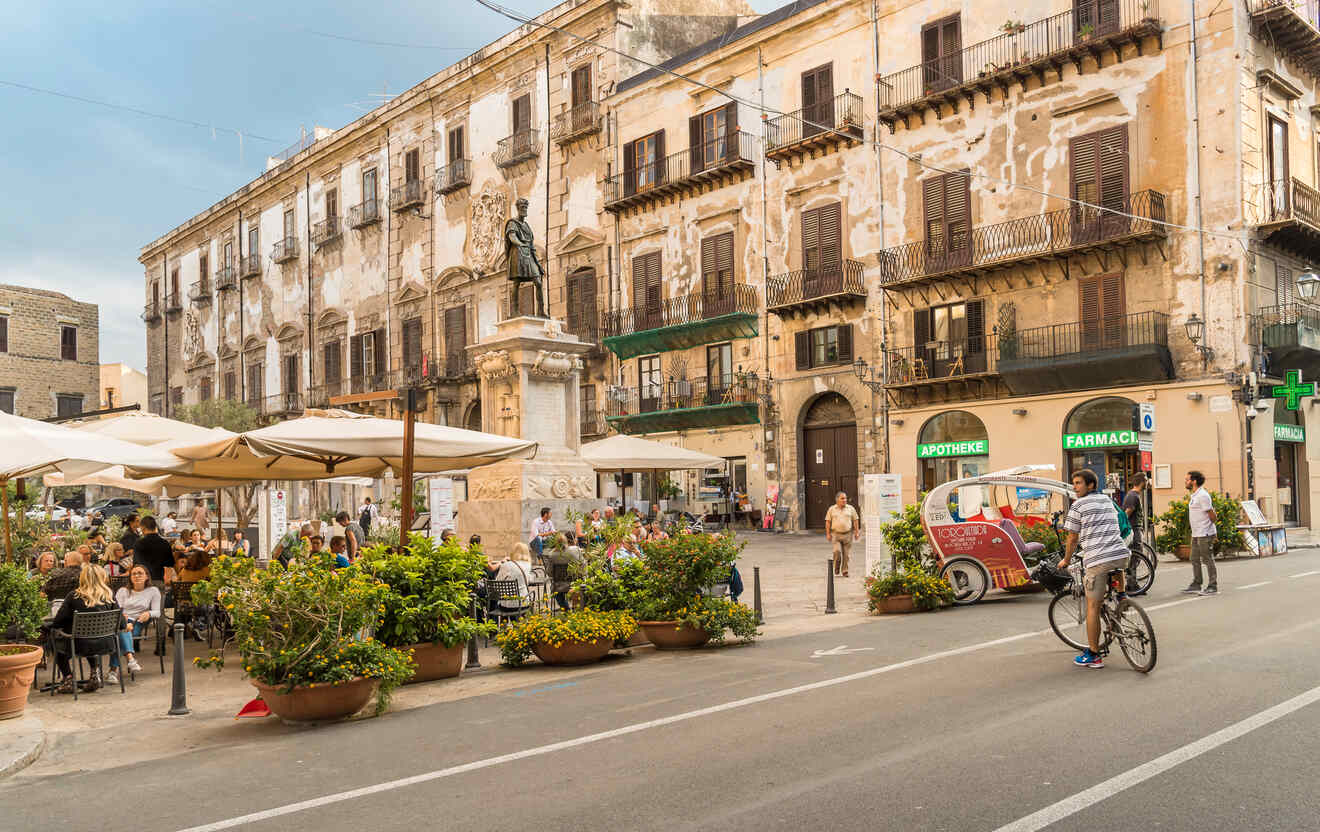 A bustling European square with outdoor cafes, people dining, an old building, and a statue in the center; a bicyclist and a colorful three-wheeled vehicle are on the road.