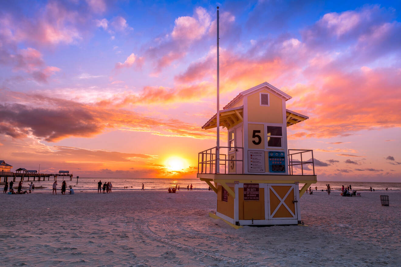 A yellow lifeguard tower with the number 5 on a sandy beach at sunset, with people in the background and a wooden pier on the left.