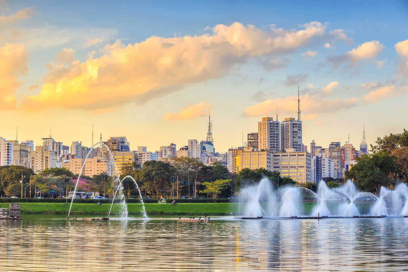 City skyline with numerous tall buildings in the background, and a park with water fountains and trees in the foreground under a partly cloudy sky at sunset.