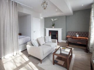 Open-plan studio at Beech House Apartments, with a cream sofa facing a fireplace, wooden coffee table, upright piano and a curtained bed area to one side.