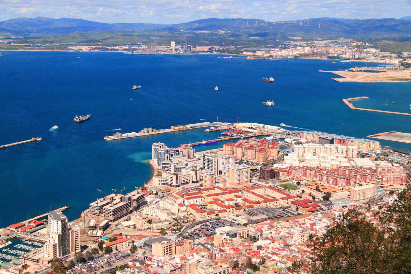 Aerial view of a coastal city with high-rise buildings, a harbor with docked ships, and a mountainous landscape in the background.