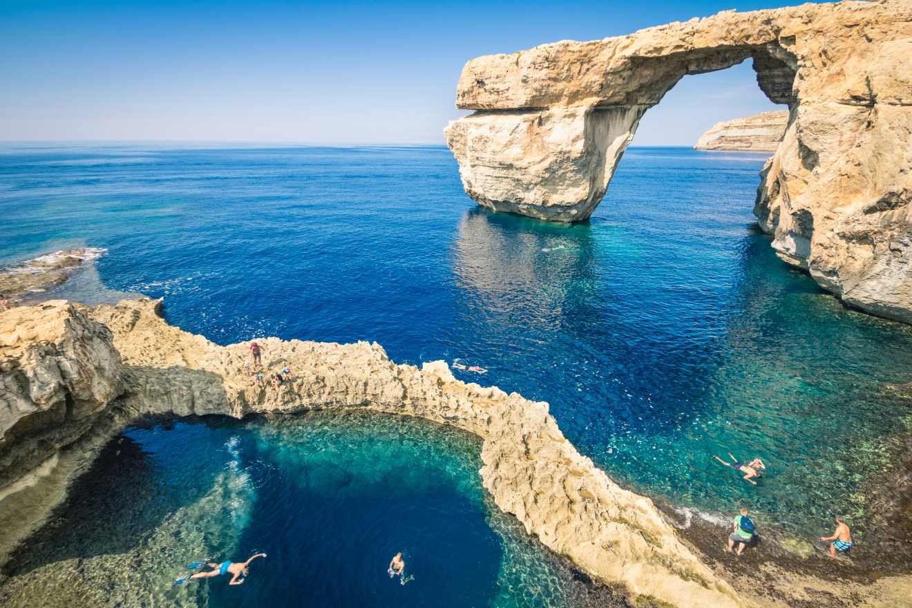 People swimming near a natural rock formation with an arch over clear blue water under a clear sky.