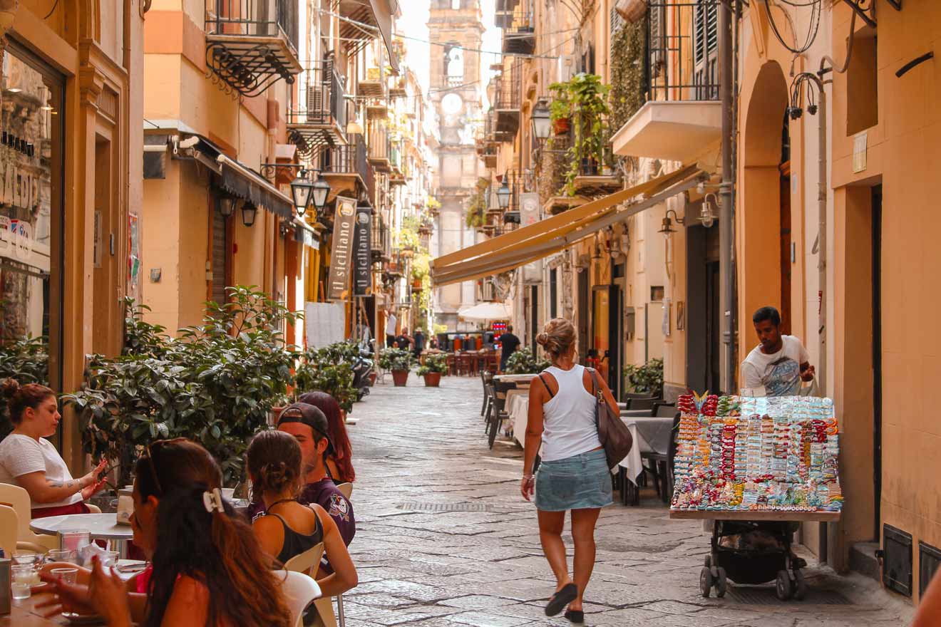 A narrow street lined with cafes and shops; people sit at outdoor tables on the left, and a person pushes a cart with goods on the right. A historical building is visible in the background.