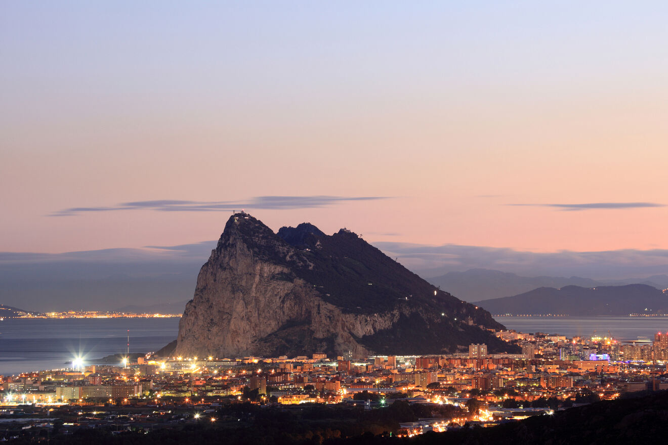 A view of the Rock of Gibraltar at sunset, with the city below illuminated by lights, and the surrounding sea and sky in soft colors.