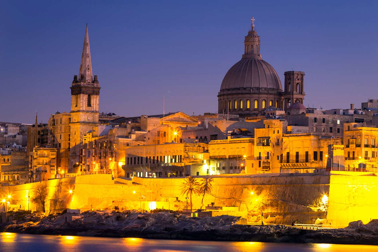 A nighttime view of Valletta, Malta, featuring illuminated historic buildings, a prominent dome, and a tall spire under a clear sky.