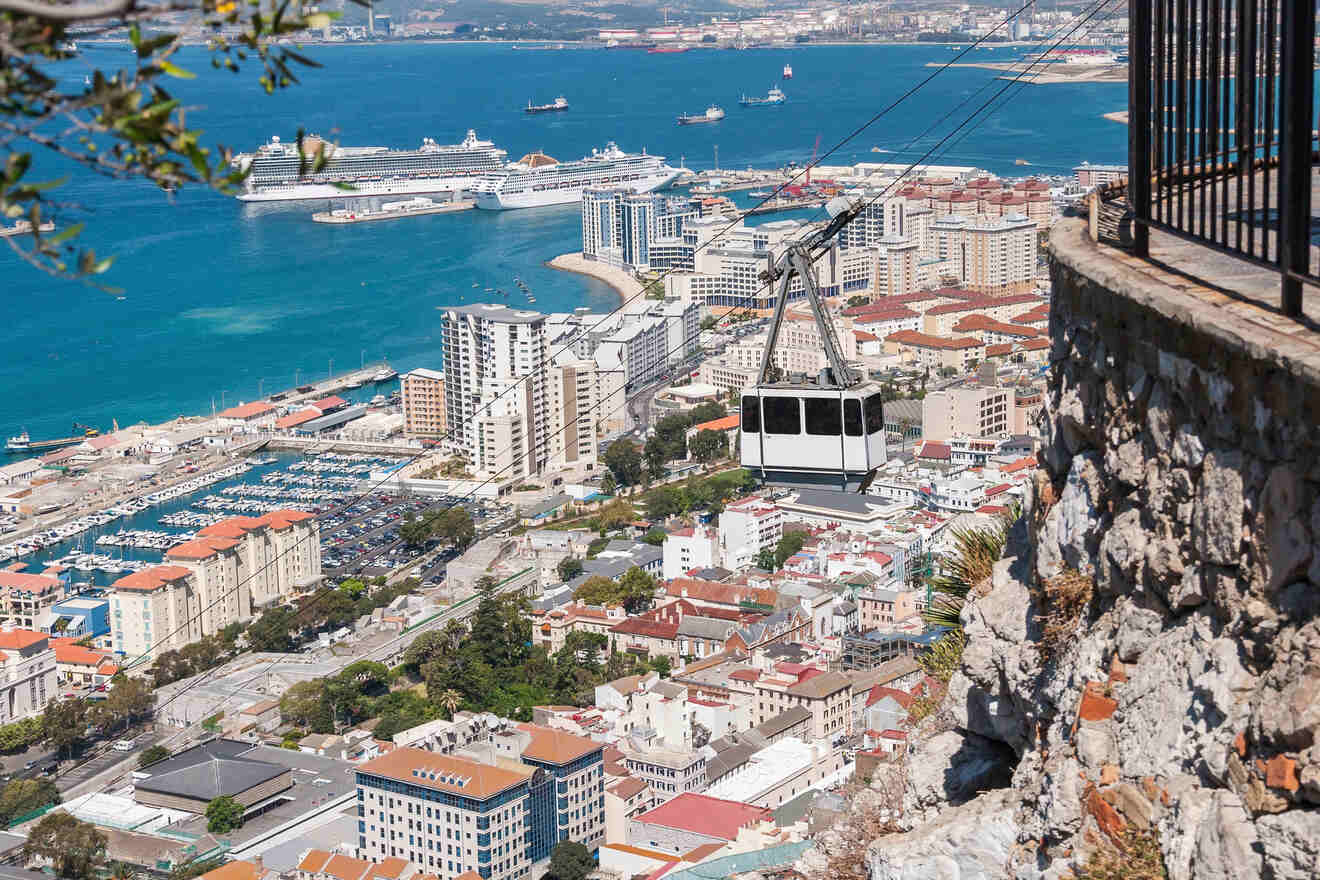 Cable car overlooking a coastal city with high-rise buildings, a harbor with cruise ships, and a rocky cliff in the foreground.