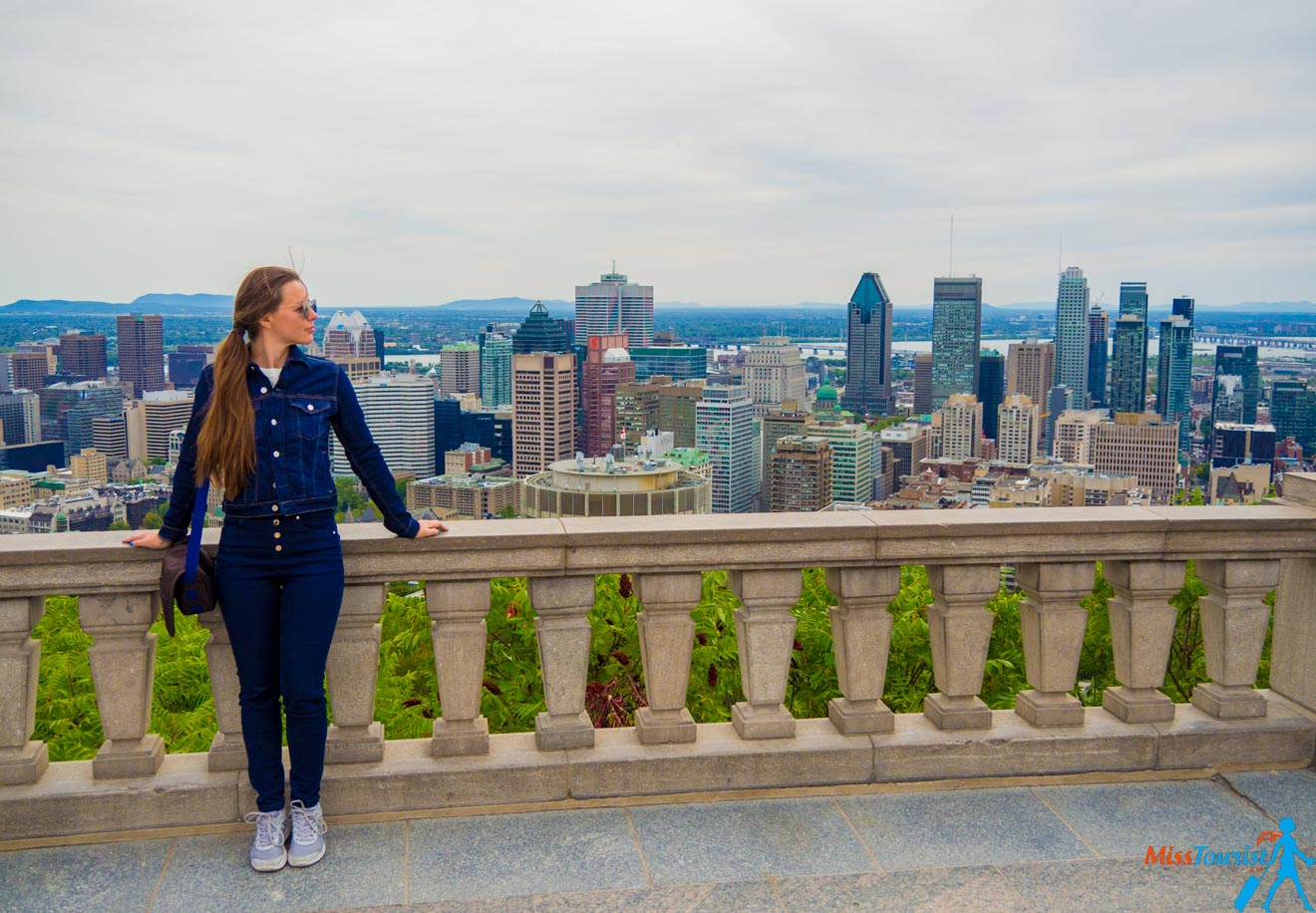 The writer of the post in a denim jacket and jeans standing at a lookout point with a panoramic view of Montreal's high-rise buildings and lush greenery in the foreground.