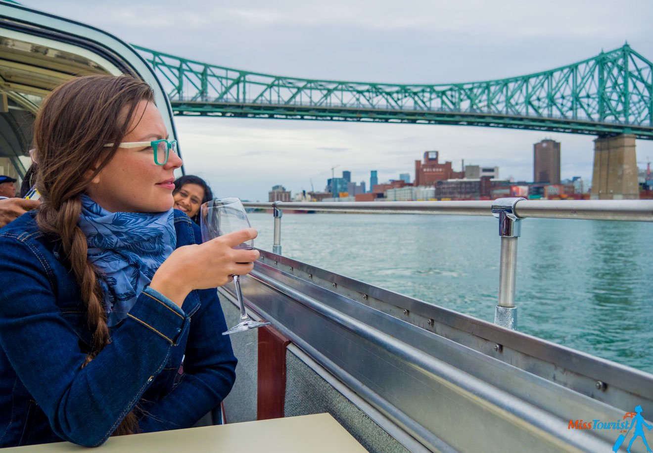 The writer of the post, Yulia, in blue attire and sunglasses holding a glass, gazing out over the water from a boat, with a green bridge and the Montreal skyline in the background
