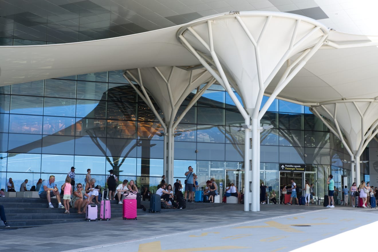 A contemporary airport terminal exterior with travelers seated and standing near the entrance under white canopy structures